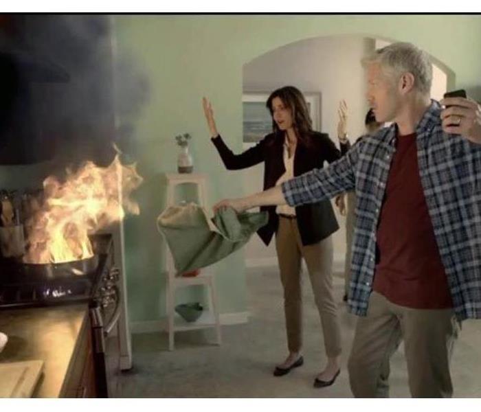 pan fire in a kitchen with man holding phone trying to put out the fire and woman with hands in the air