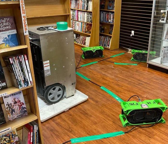 green air movers and a silver dehumidifier sitting on a brown wood floor in a comic book store